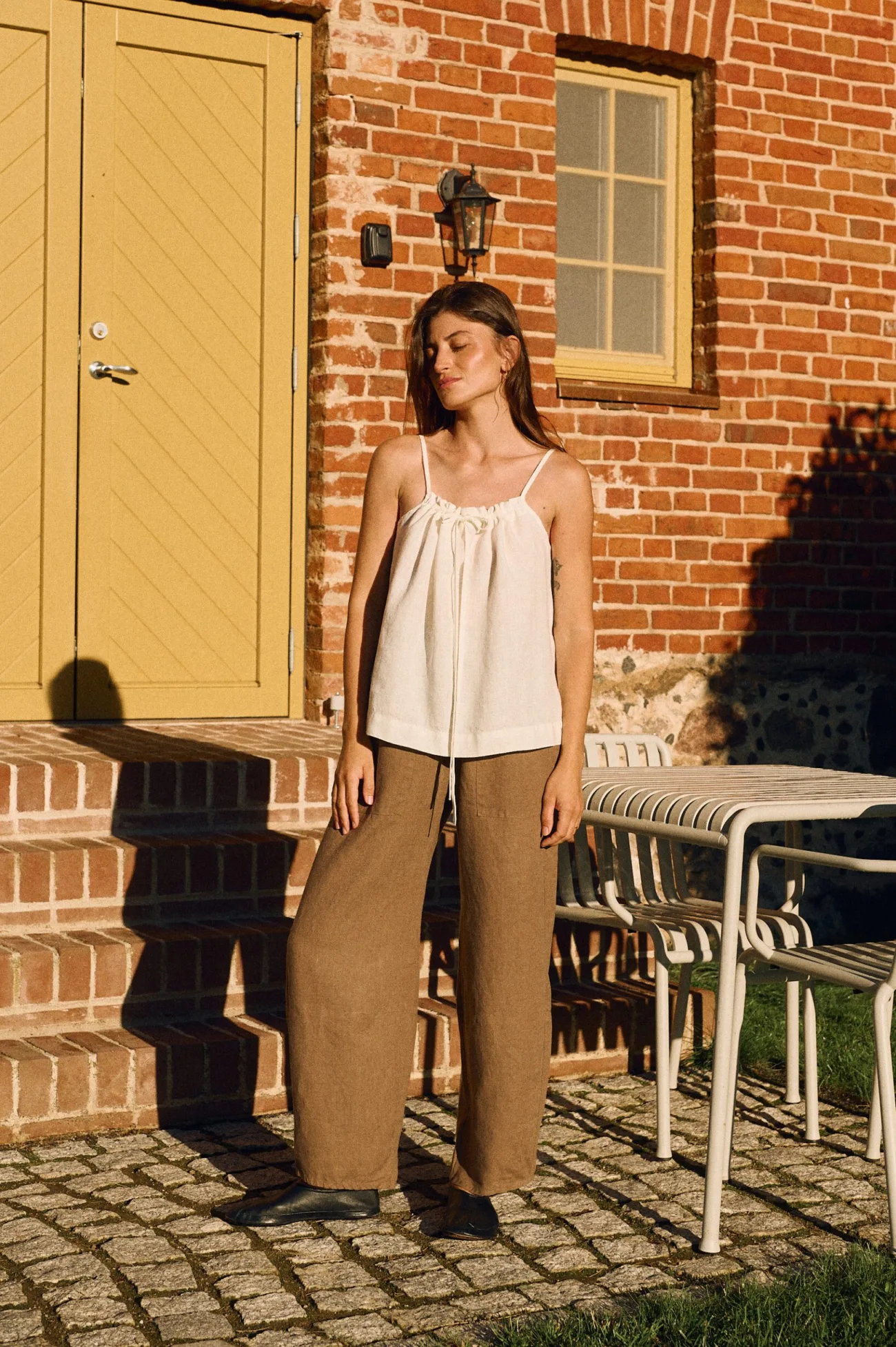 Minimalist white linen camisole top with adjustable drawstring front and thin straps, styled with wide-leg linen trousers