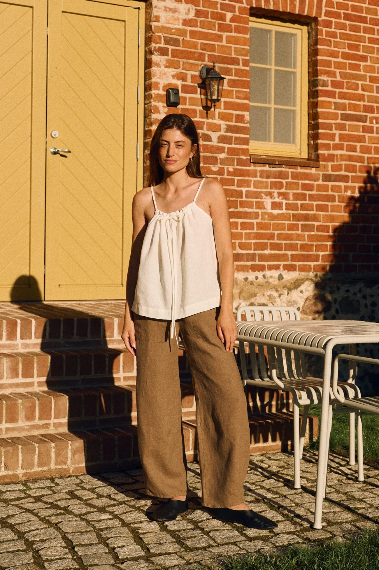 Model wearing a white linen camisole top with adjustable drawstring neckline paired with cacao brown wide-leg linen trousers