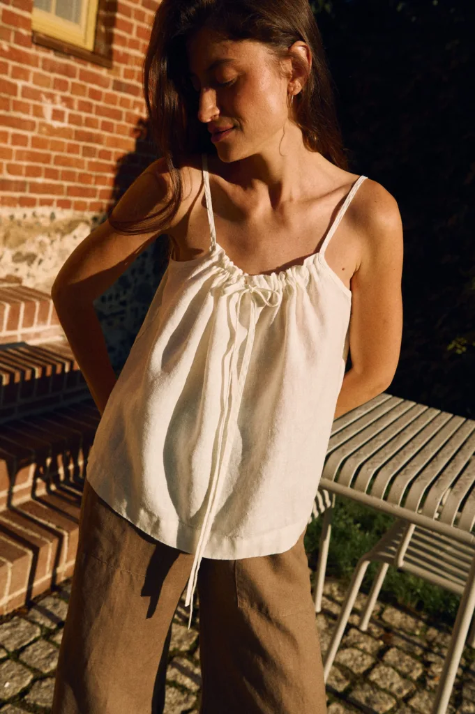 White linen camisole top with adjustable drawstring neckline and thin straps, made from breathable natural linen fabric, styled with wide-leg linen trousers