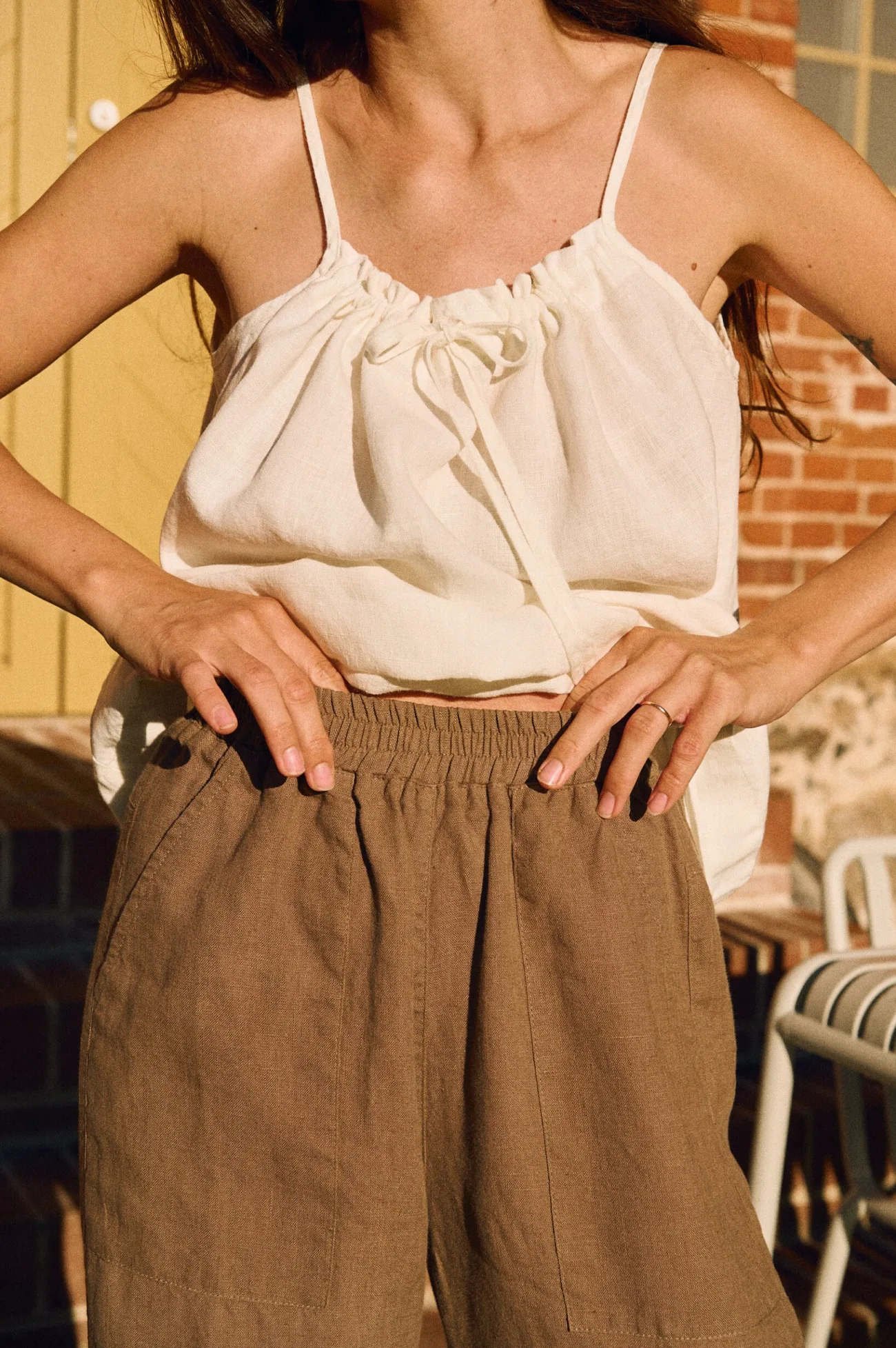 Close-up of cacao linen trousers showing elasticated waist and deep patch pockets, paired with milky white linen cami with gathered neckline