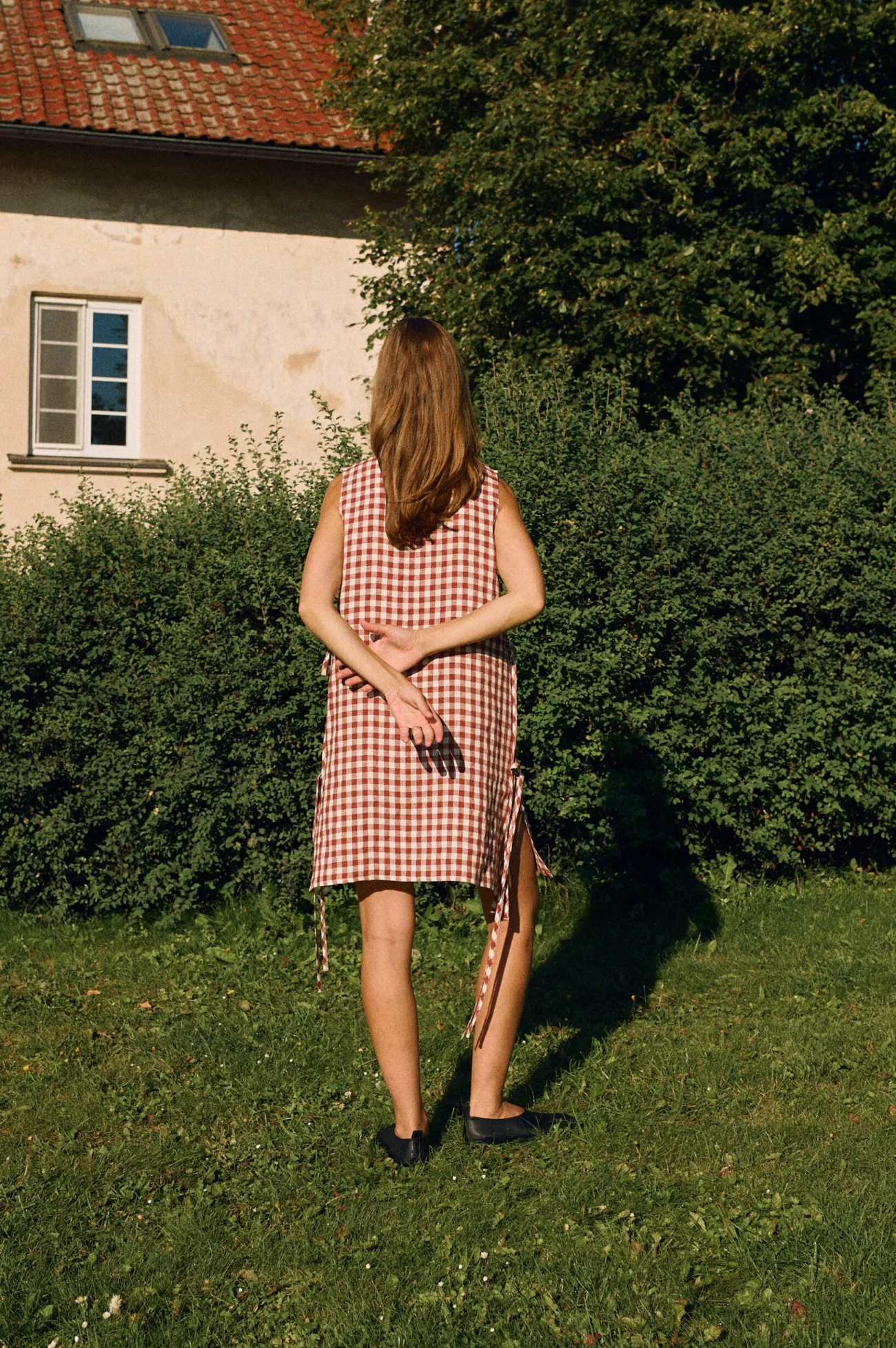 Model seen from the back wearing a sleeveless midi dress in terracotta gingham linen with side ties