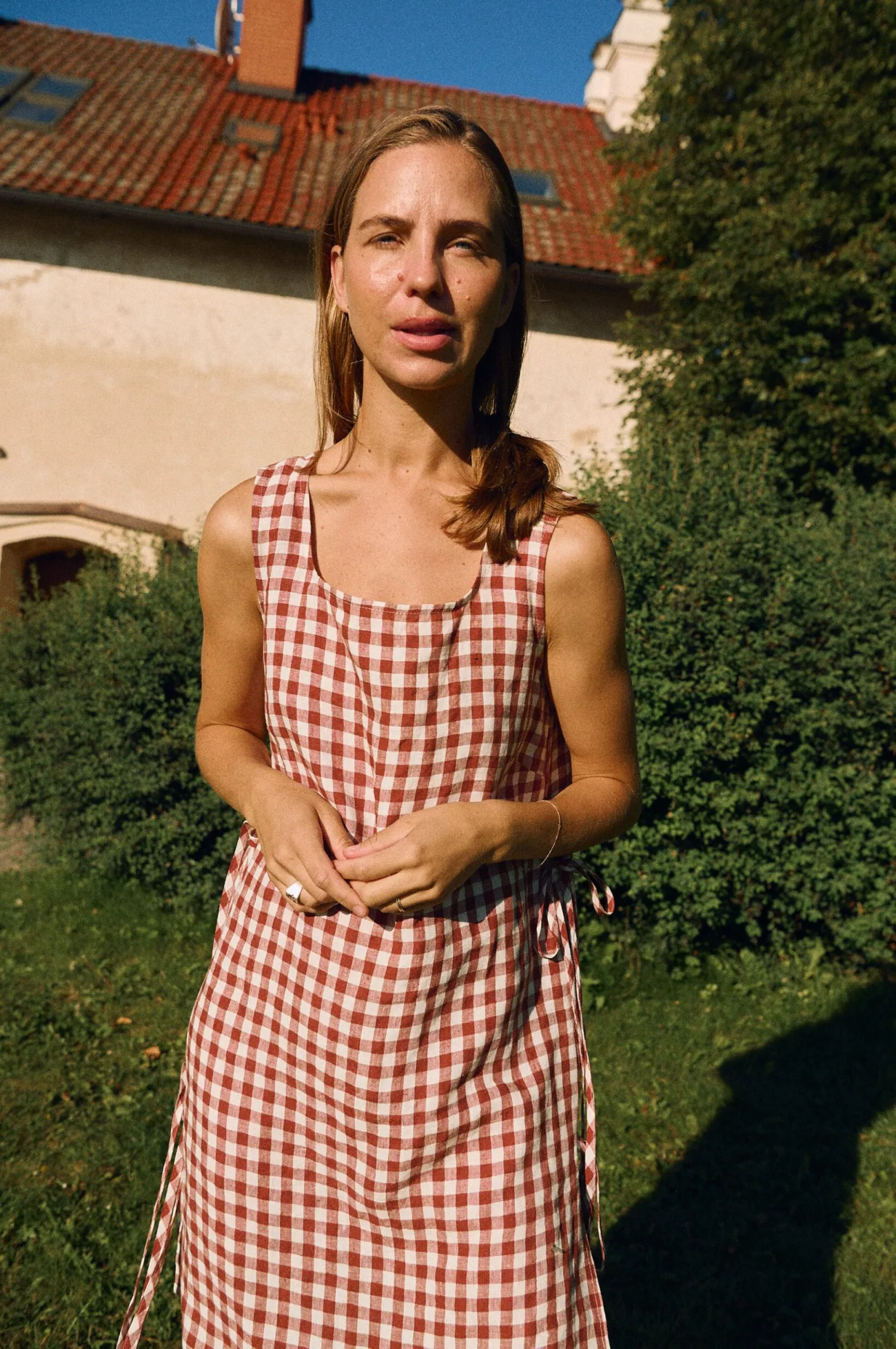 Woman wearing a sleeveless terracotta and white gingham linen midi dress with a squared scoop neckline, side tie details, and side slits