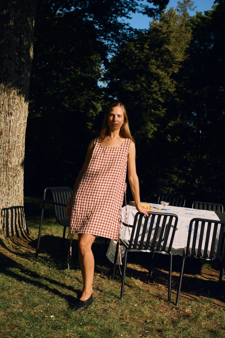 Woman wearing a sleeveless dress in terracotta gingham linen, featuring a square neckline, bust darts for gentle bust shaping, side ties, and a relaxed, breathable silhouette with a midi-length hem