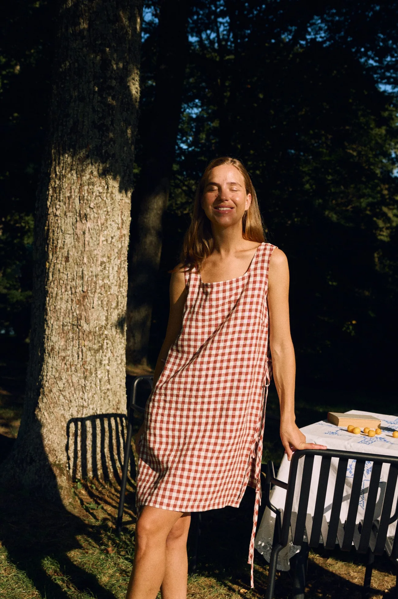 Woman wearing the sleeveless Ella dress in terracotta gingham linen, featuring a square neckline, chest darts, side ties, and a relaxed midi-length silhouette