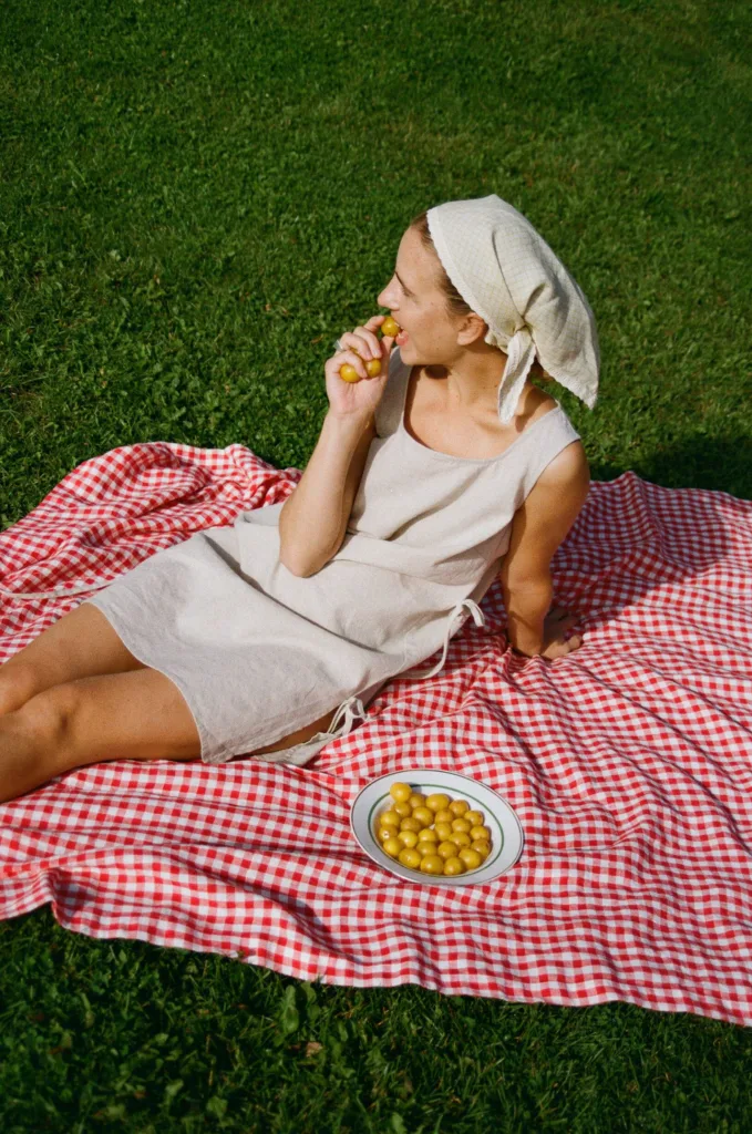 Woman wearing natural sleeveless cotton-linen blend dress with side tie details and apple checks linen headscarf lying on red gingham blanket outdoors