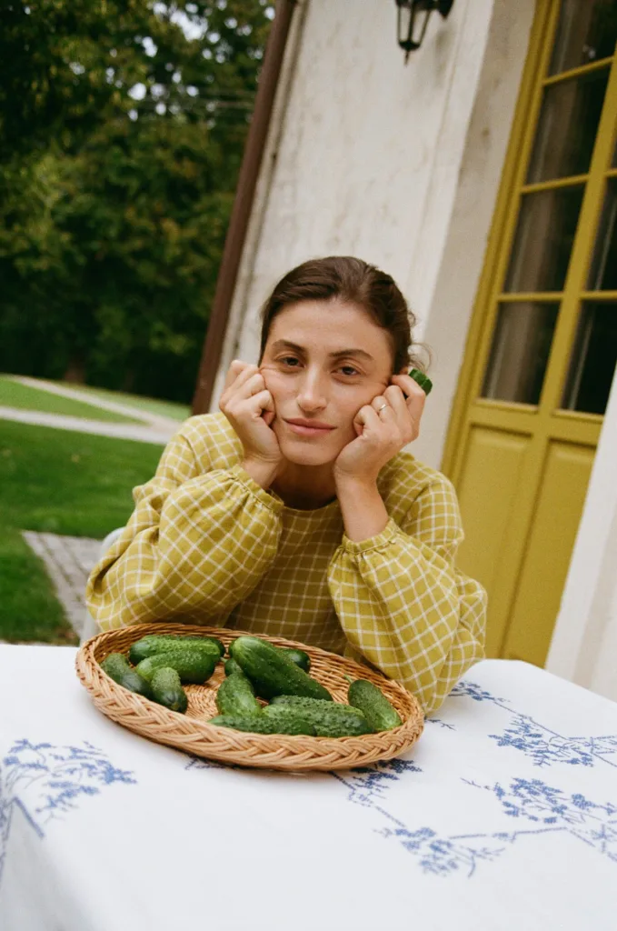 Woman wearing pickle checks linen dress seated at table with basket of cucumbers