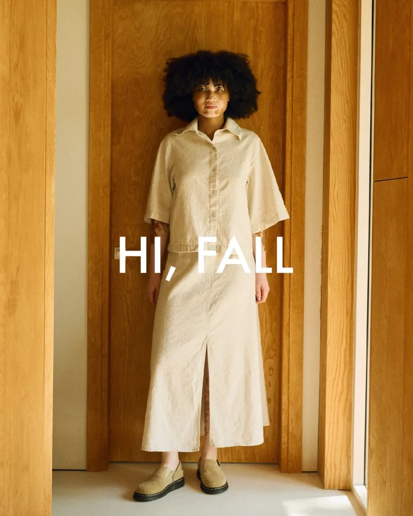 Model wearing a natural linen-cotton blend top with front snaps and wide sleeves, paired with a matching long skirt featuring a front slit, standing in a wooden interior with the text “HI, FALL” overlaid
