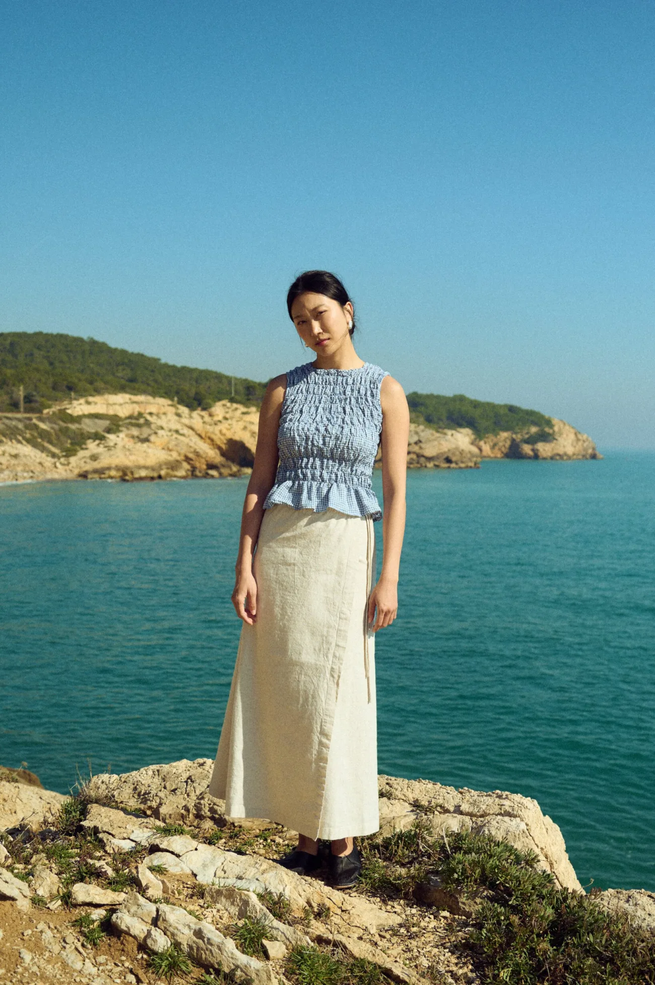 Front view of a white cotton-blend maxi skirt paired with a dusty blue mini gingham linen sleeveless top with a smocked bodice and peplum hem