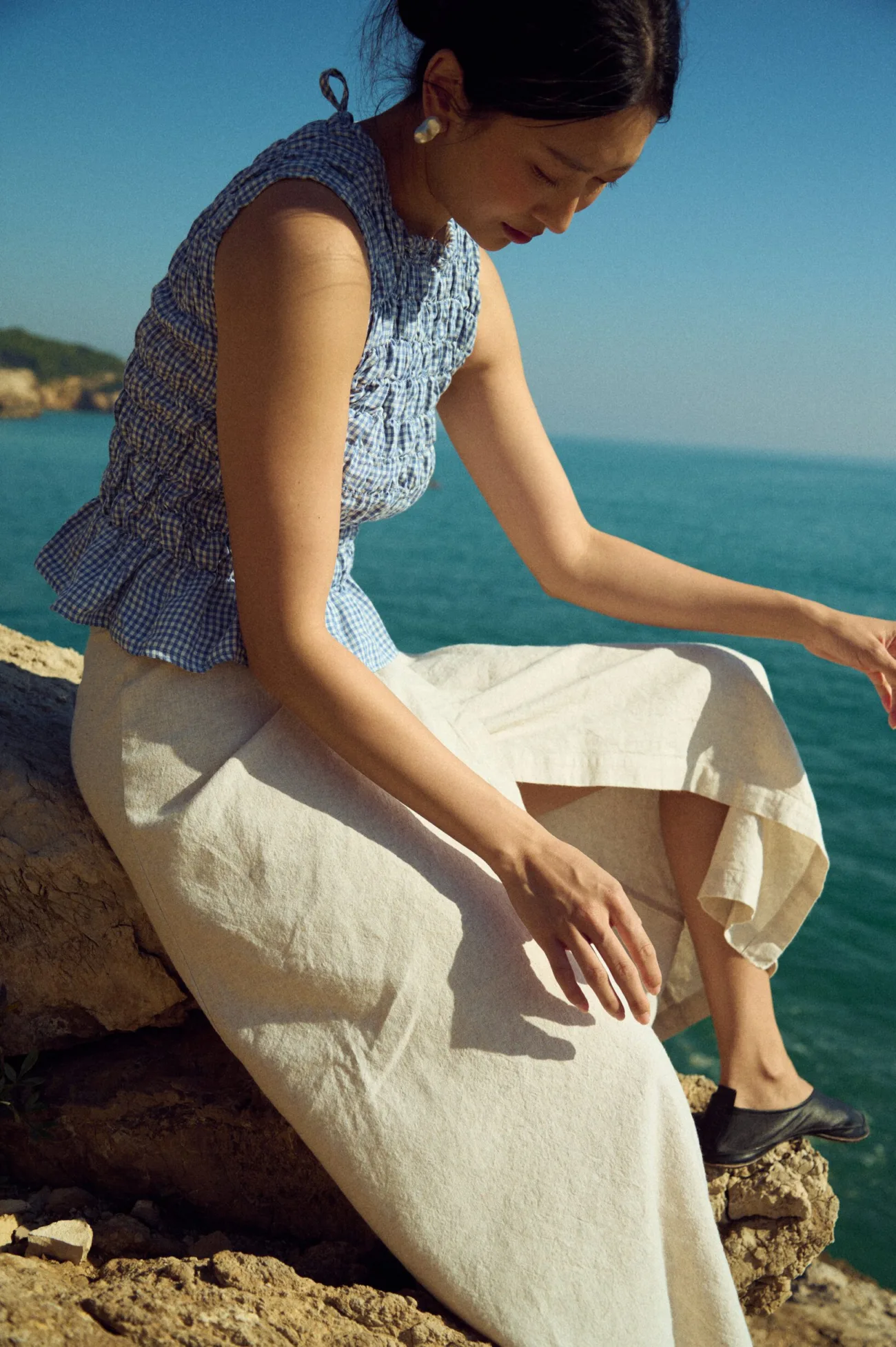 Seated view of a white cotton-blend wrap maxi skirt with tie detail, paired with a blue mini gingham linen sleeveless smocked top