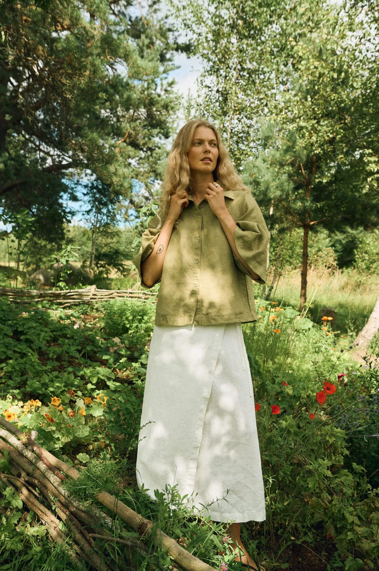 Woman wearing an olive green linen shirt jacket with snap button closure and wide sleeves, styled with a milky white linen wrap skirt featuring a tie waist