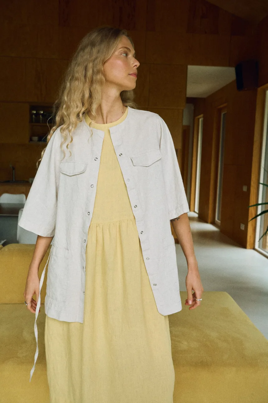 Light yellow sleeveless linen midi dress styled with an open-front natural linen wool blend overshirt featuring half sleeves, flap chest pockets, and snap button closure