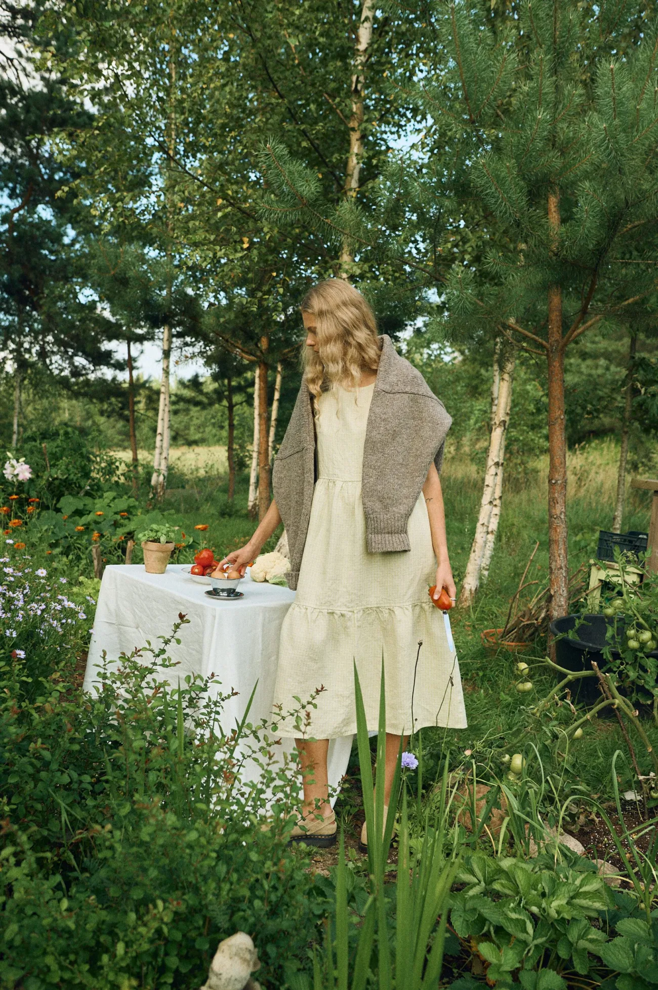 Sleeveless tiered dress in apple checks linen styled with a chunky knit sweater draped over the shoulders, featuring a relaxed silhouette and round neckline
