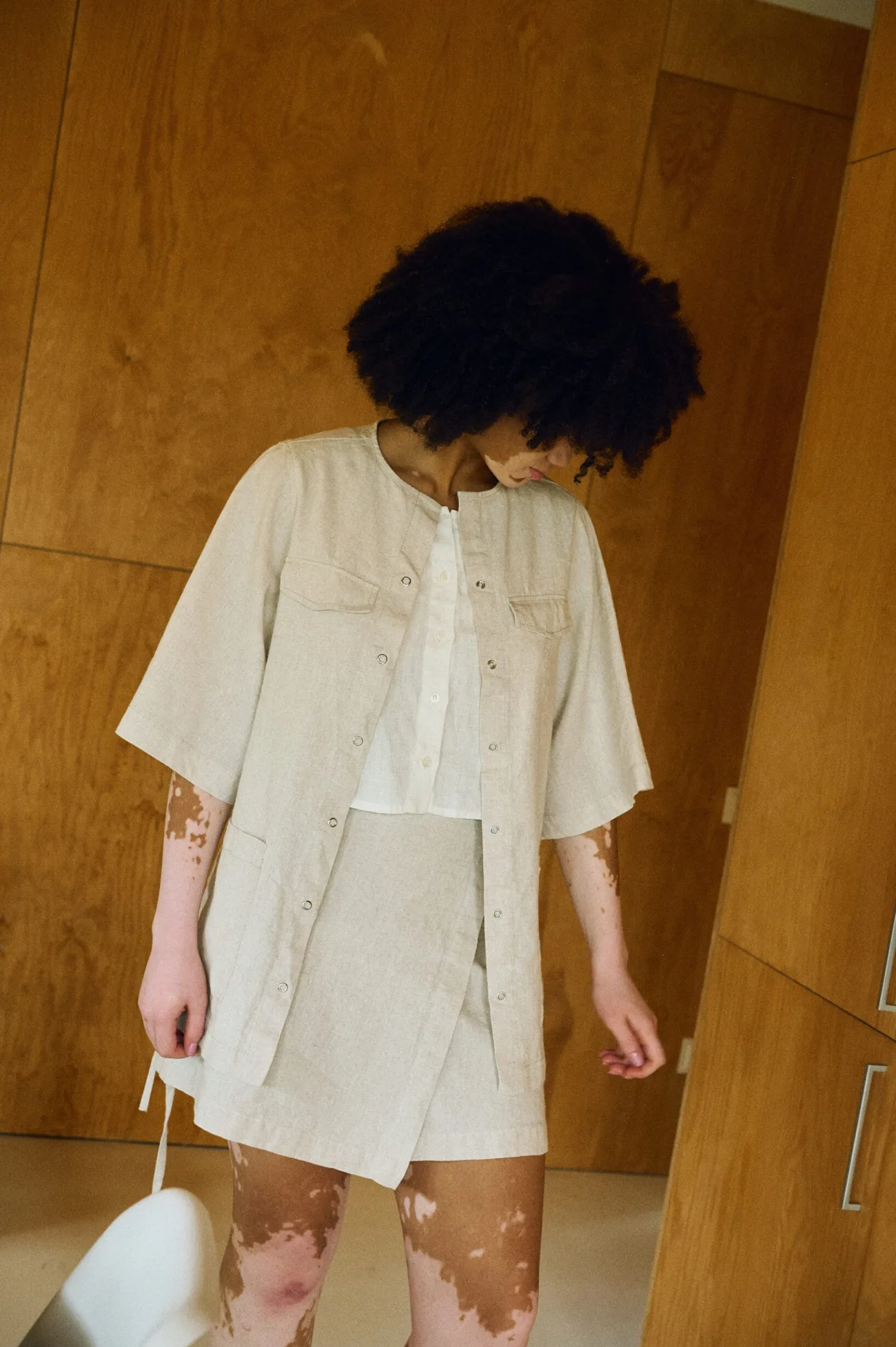 Downward-facing view of a model wearing a natural cotton blend overshirt with snap closures and wide sleeves, layered over a cropped milky white linen top and paired with a matching natural cotton blend wrap-style skirt