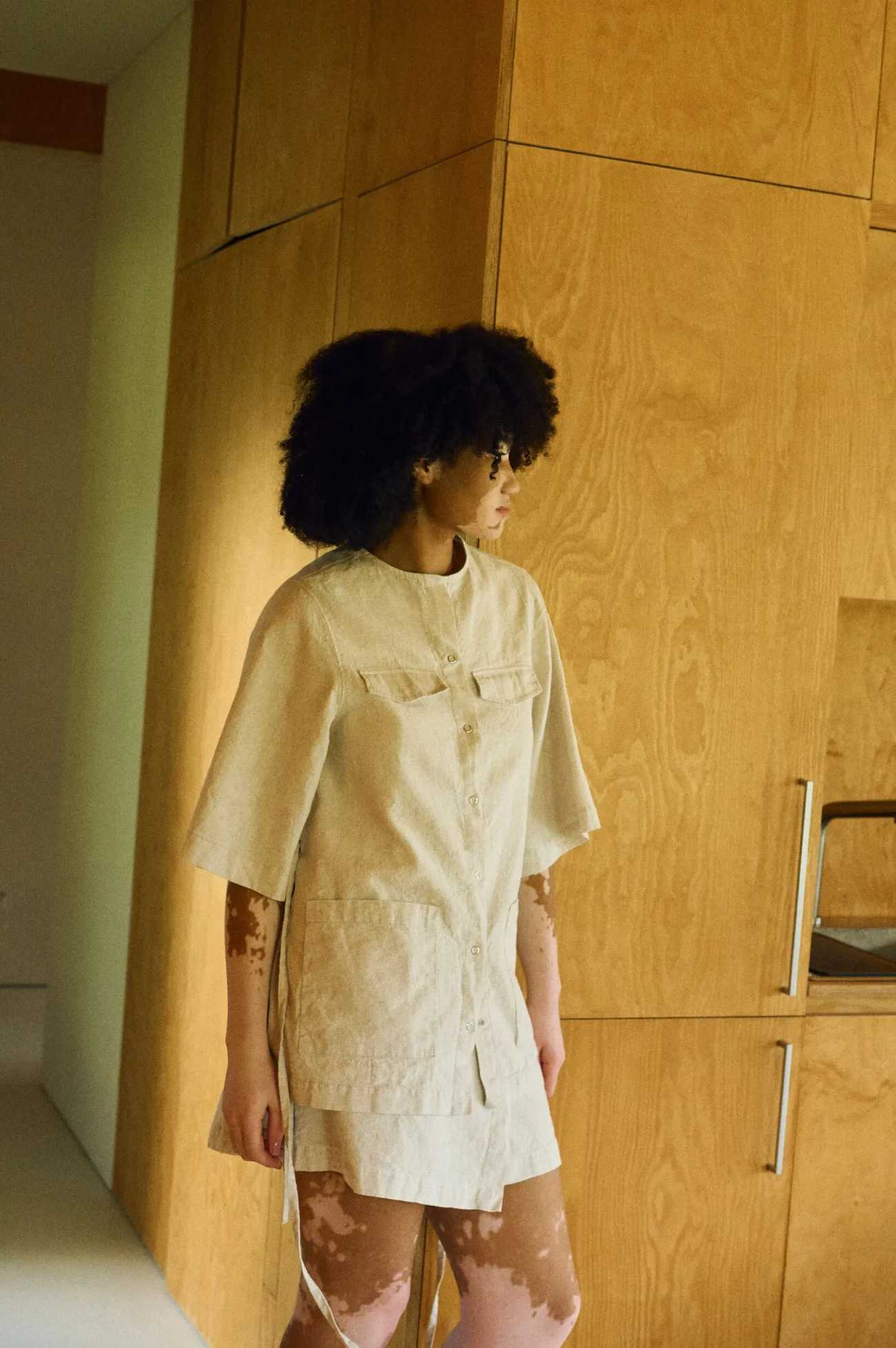 Side view of a model wearing a natural cotton blend overshirt with snap closures, chest flaps, and wide elbow-length sleeves, paired with a matching wrap-style mini skirt in the same fabric