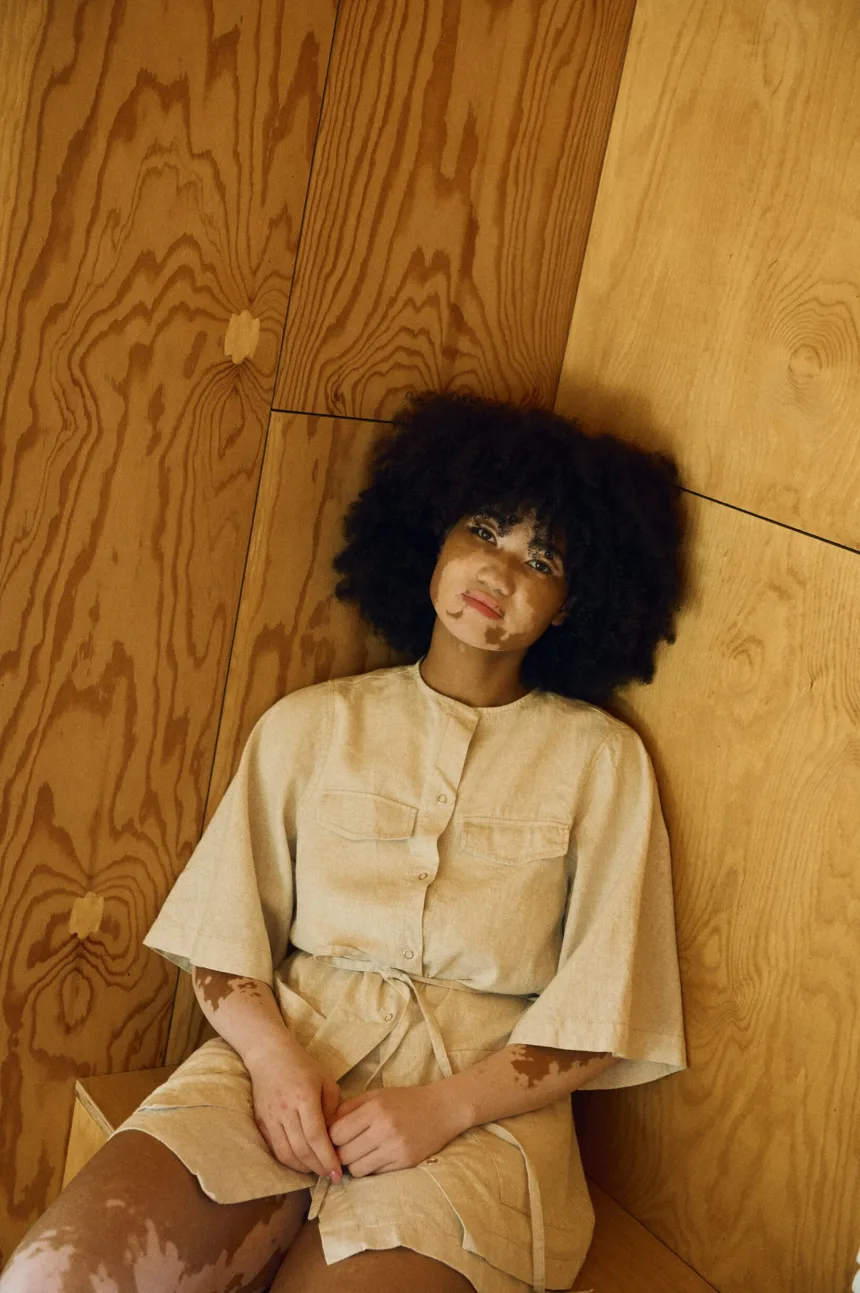 Model seated wearing a natural cotton blend overshirt with snap closures, chest flaps, and wide elbow-length sleeves, styled as a belted dress over a matching wrap-style skirt