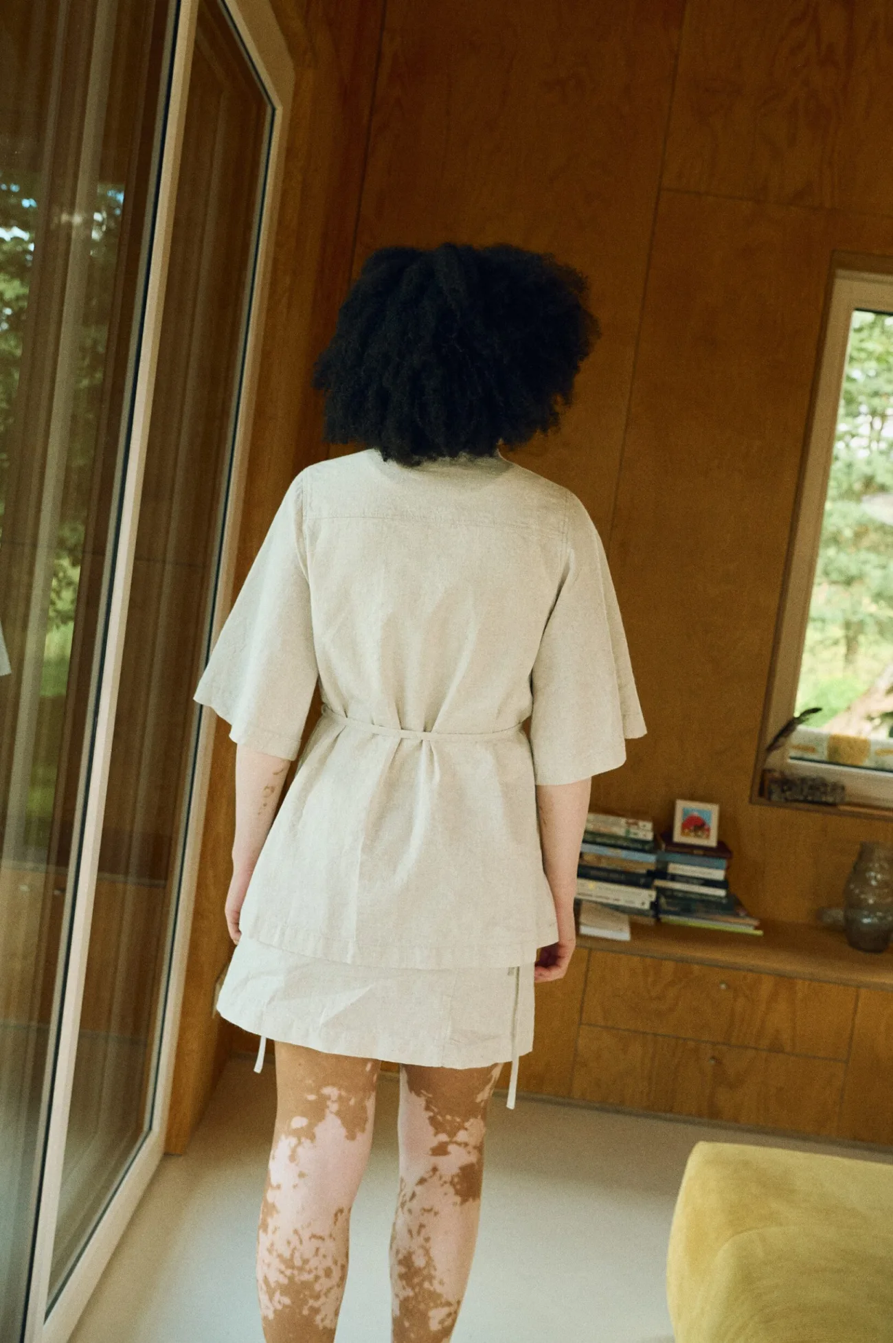 Back view of a model wearing a natural cotton blend overshirt cinched at the waist with a tie belt, paired with a matching wrap-style mini skirt
