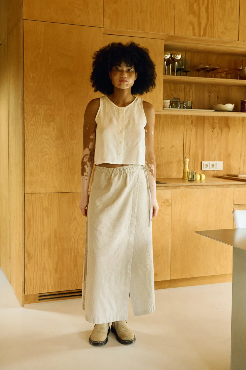 Woman wearing a natural cotton-linen blend ankle-length wrap skirt with a clean A-line silhouette, flat front, and long adjustable ties, paired with a sleeveless button-up top