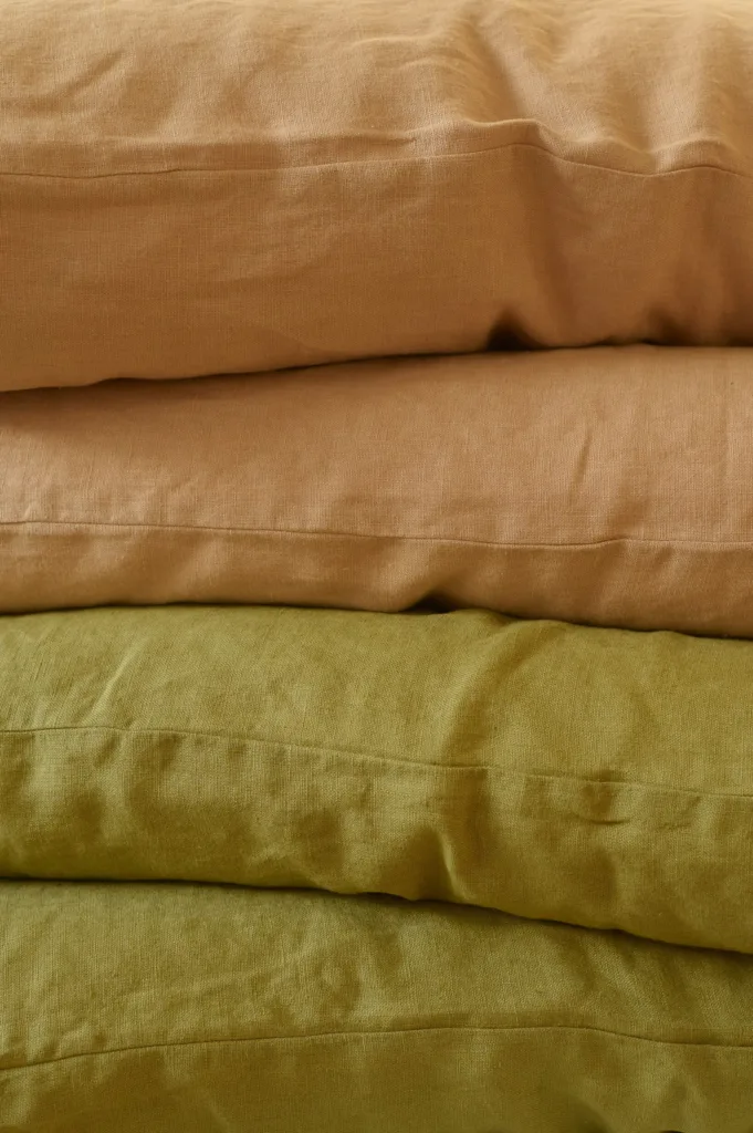 Stack of softened linen pillowcases in warm earthy tones, including dusty peach and olive green, showcasing the natural texture and relaxed drape of high-quality linen bedding