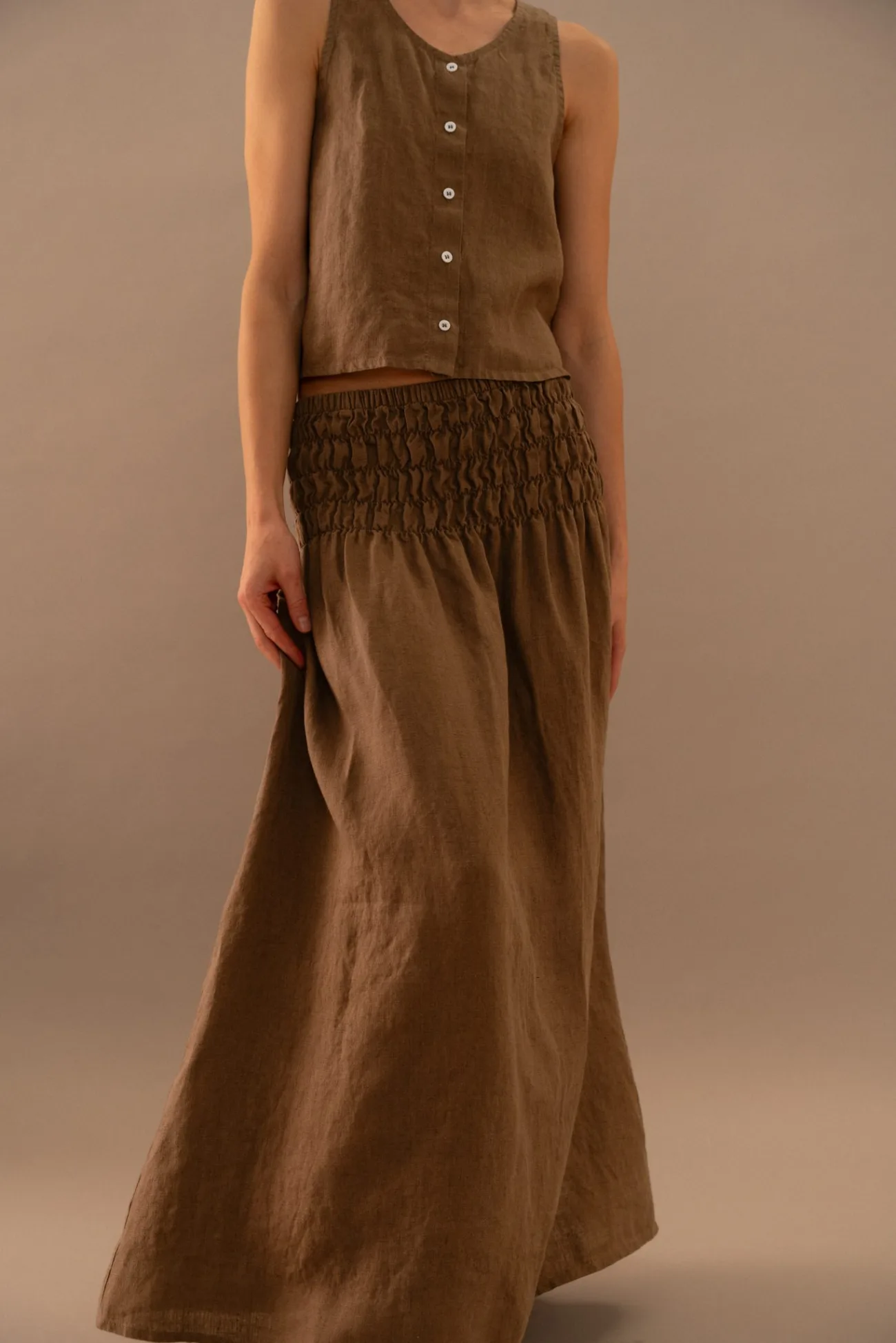 Model wearing cacao linen two piece set with sleeveless button front top and flowing maxi skirt in soft neutral studio light