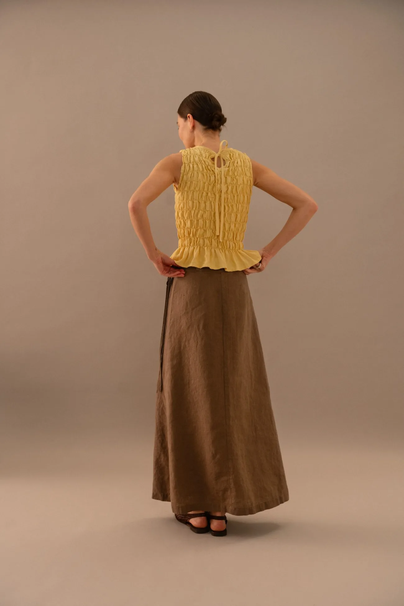 Back view of butter yellow sleeveless smocked linen top with ruffled hem and tie closure, styled with cacao brown linen wrap maxi skirt