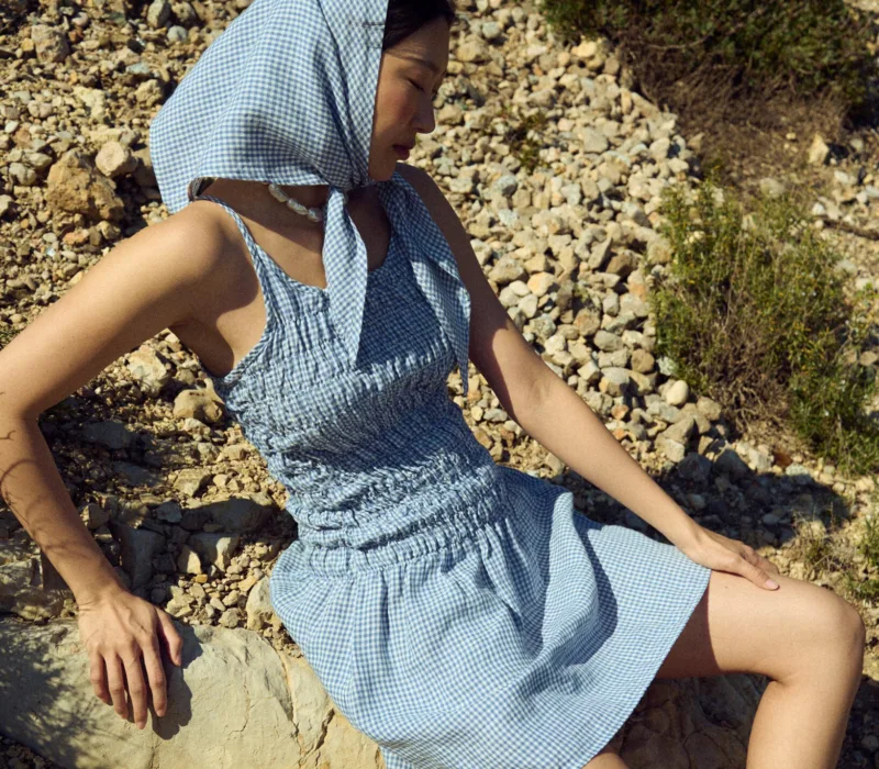 Seated side view of a dusty blue mini gingham linen mini dress with thin straps and a ruched smocked bodice, styled with a matching headscarf