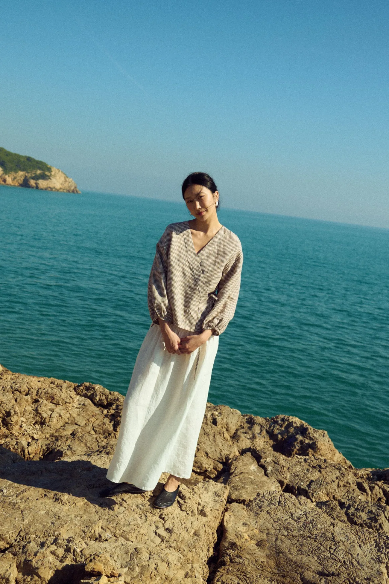 Front view of a milky white linen maxi skirt with a smocked elastic waistband paired with a brown mini gingham wrap blouse with tie closure