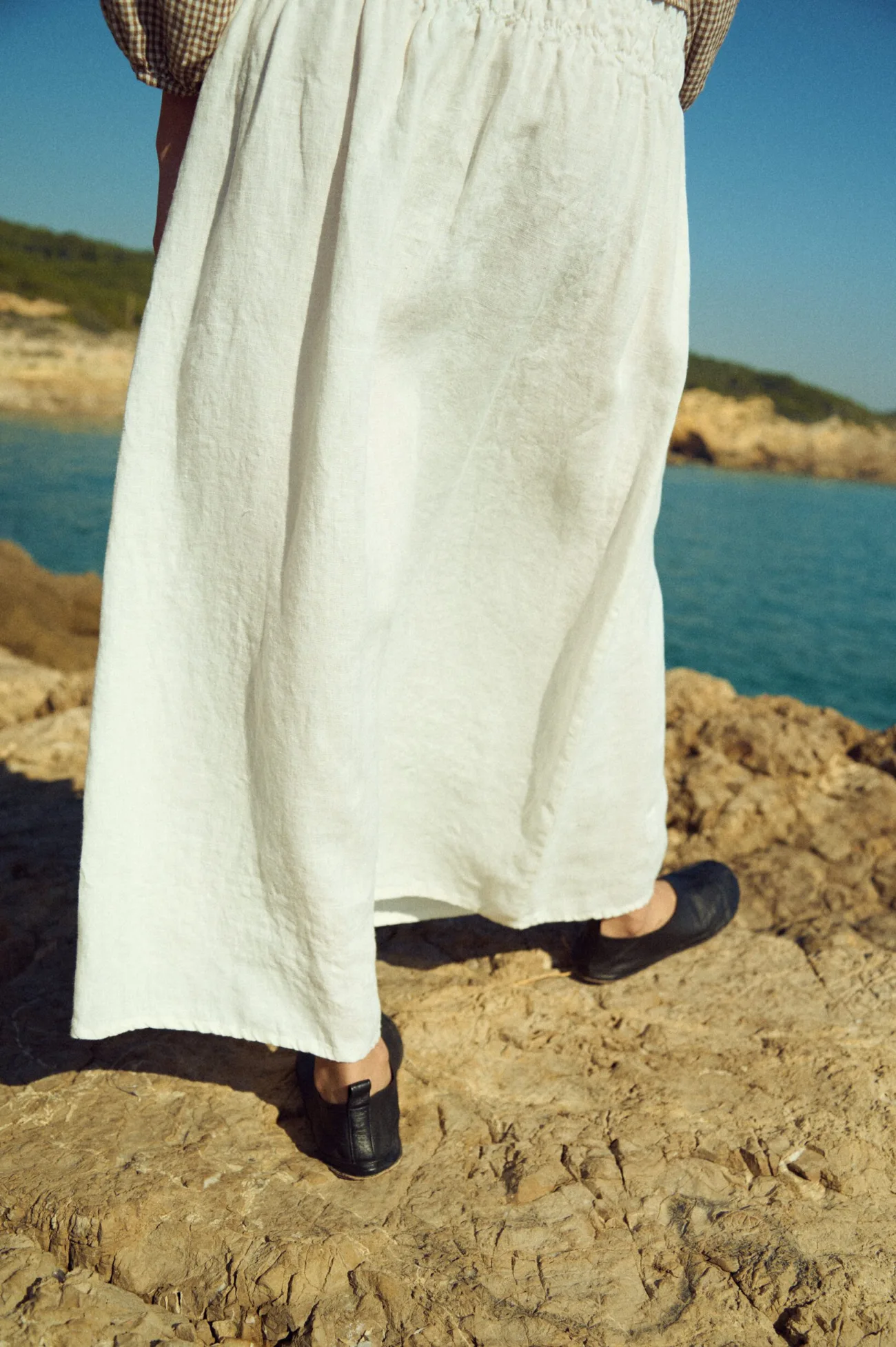 Back view of a milky white linen maxi skirt with a wide smocked elastic waistband and softly draped, flowy hem