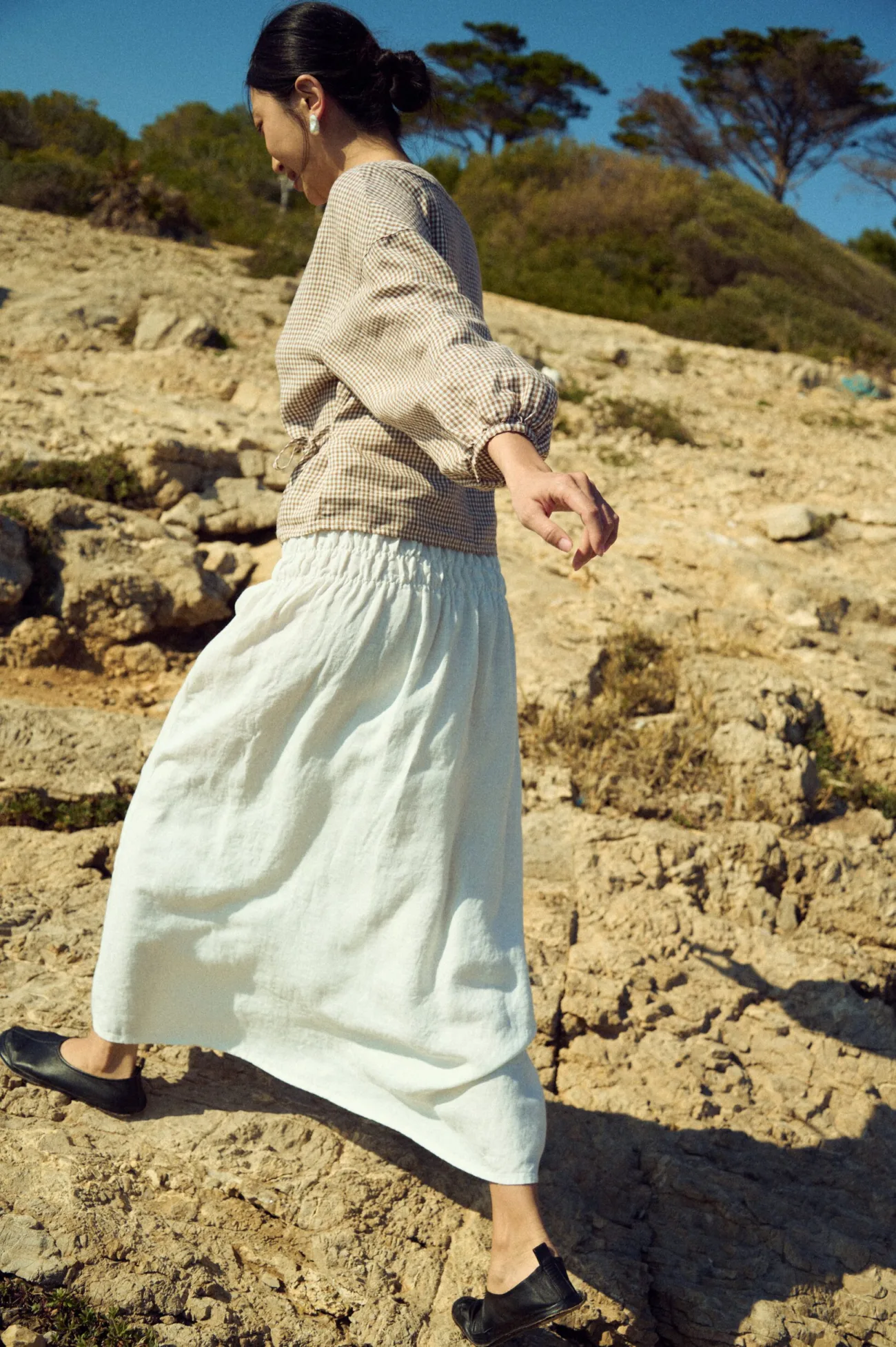 Side view of a milky white linen maxi skirt with a smocked elastic waistband paired with a brown mini gingham blouse with gathered sleeves