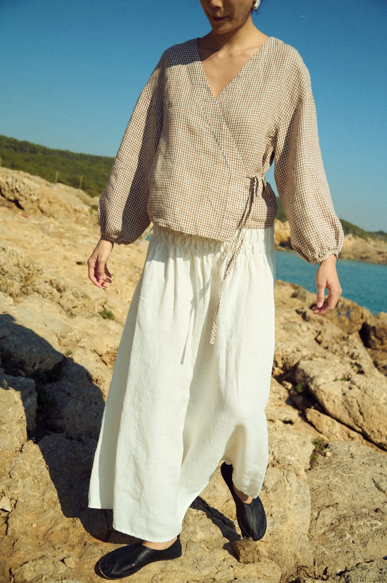 Front view of an off-white linen maxi skirt with a smocked elastic waistband paired with a brown mini gingham wrap blouse with tie detail