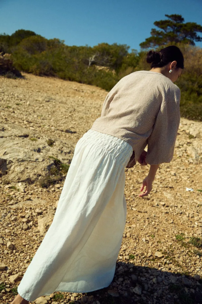 Back view of a milky white linen maxi skirt with a smocked elastic waistband paired with a brown mini gingham blouse with relaxed sleeves