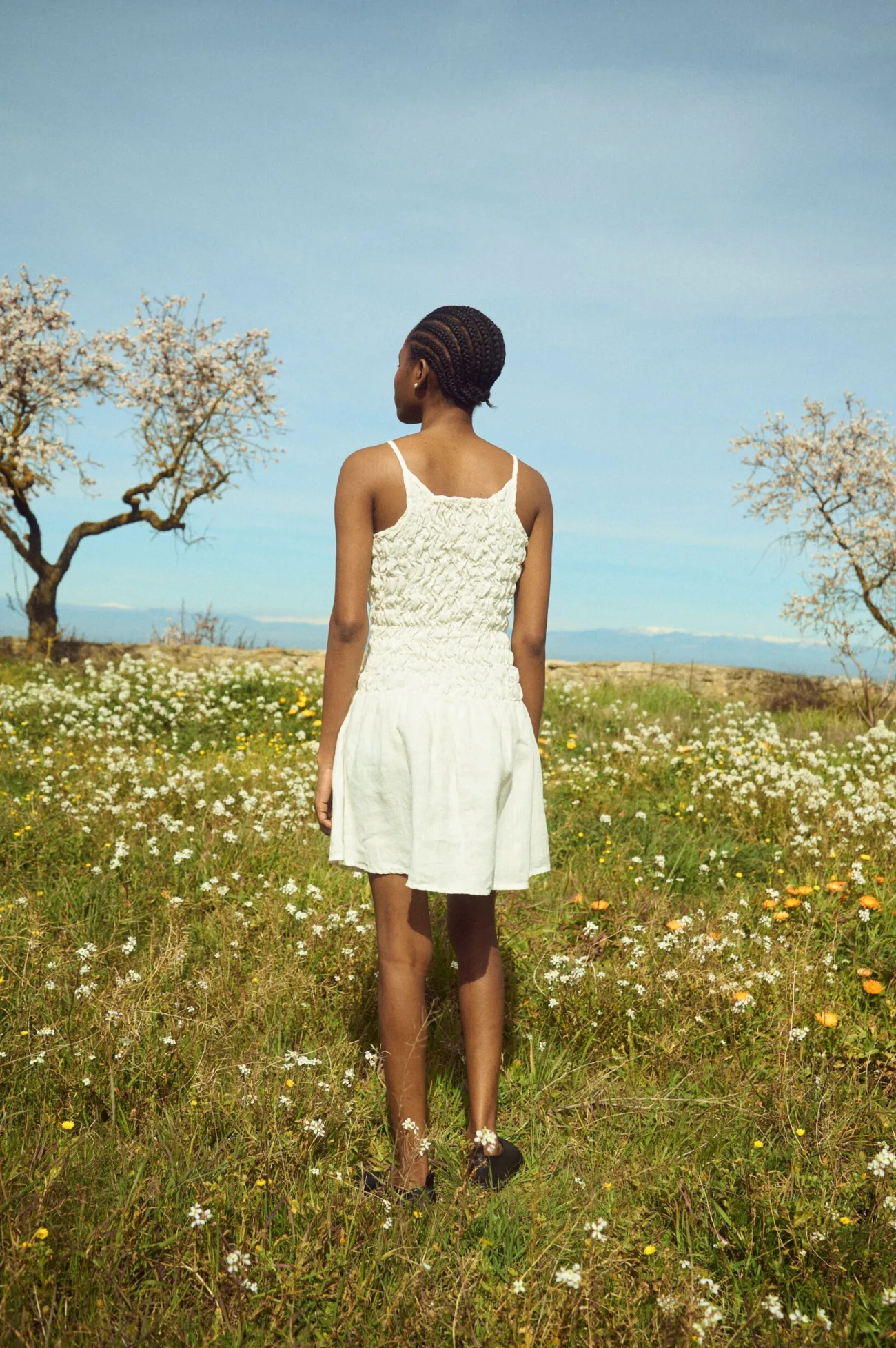Back view of a milky white linen mini dress with thin straps, a ruched smocked bodice, and a softly gathered skirt