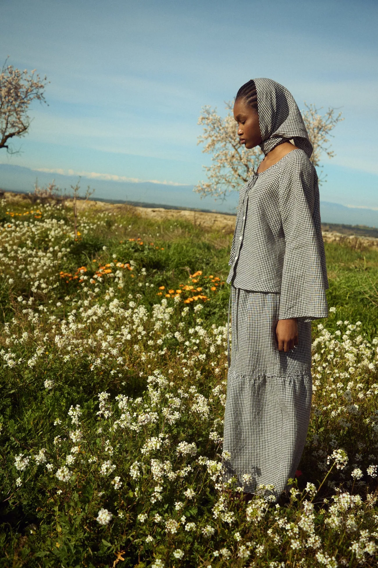 Profile view of a black mini gingham linen blouse with wide sleeves and a matching tiered gingham skirt, styled with a coordinating headscarf