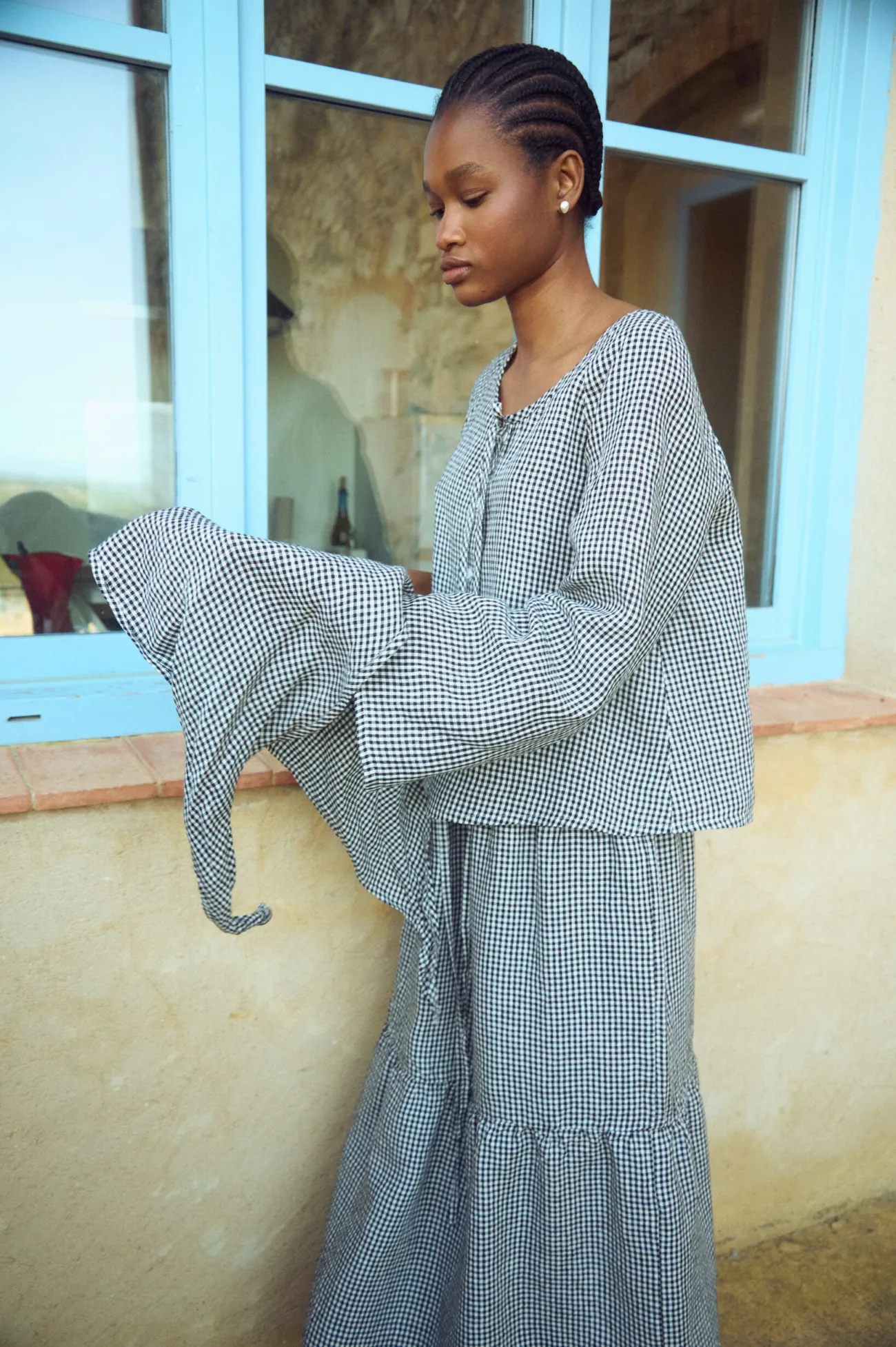 Side view of a black mini gingham linen blouse with voluminous sleeves and a matching tiered gingham skirt, holding a coordinating headscarf
