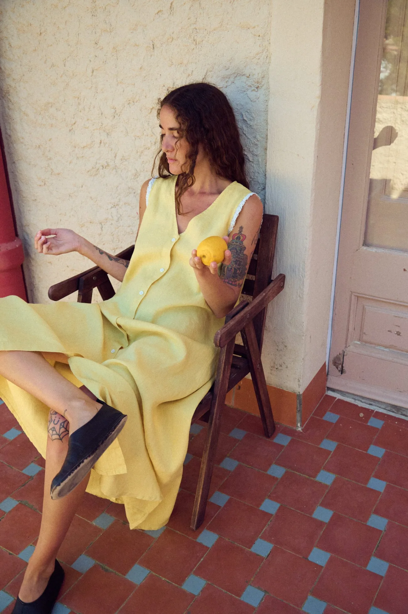 Seated woman in a yellow linen sleeveless maxi dress with V-neck, button front, lace-trimmed armholes, and fluid A-line shape