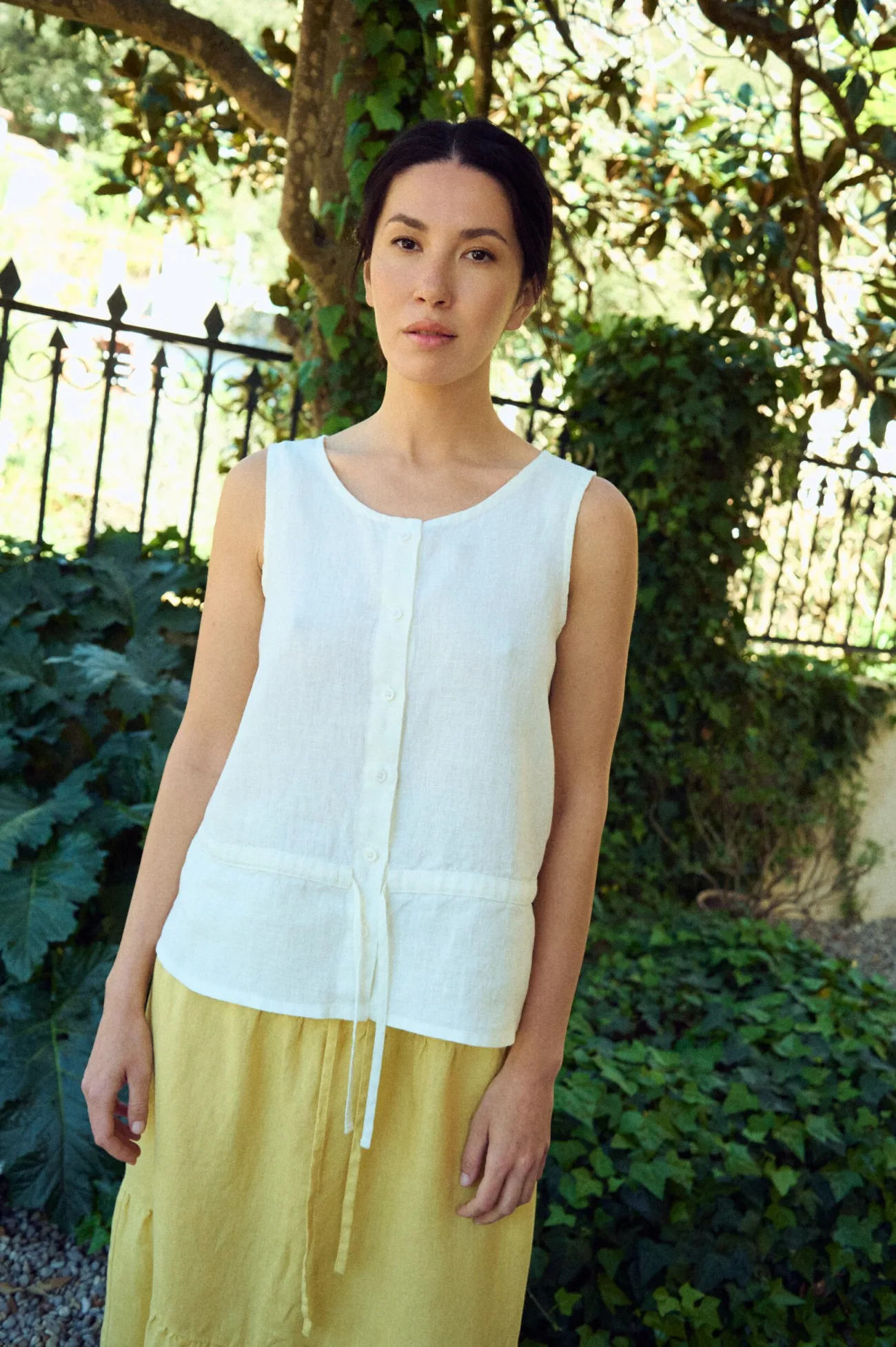 White sleeveless linen top with a relaxed fit, round neckline, button-front closure, and subtle waist seam with tie detail