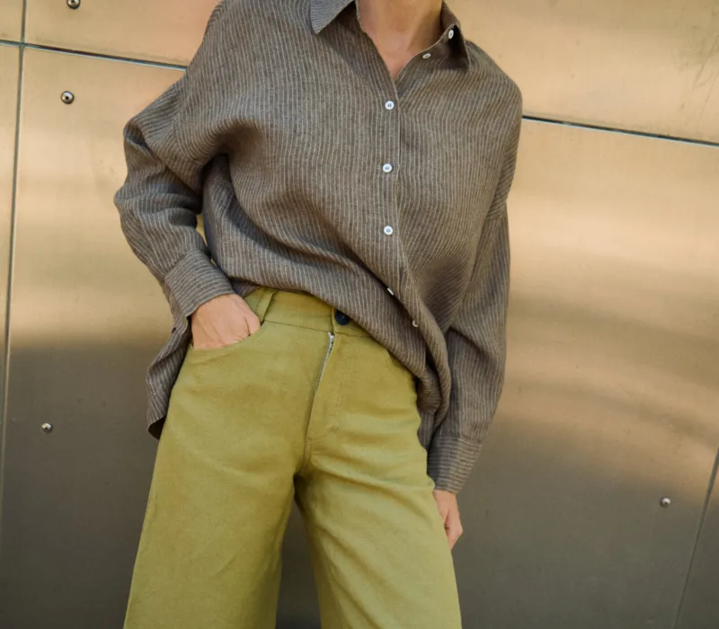 Close-up of olive heavy linen pants showing the front button closure and curved side pocket, styled with a brown striped linen shirt