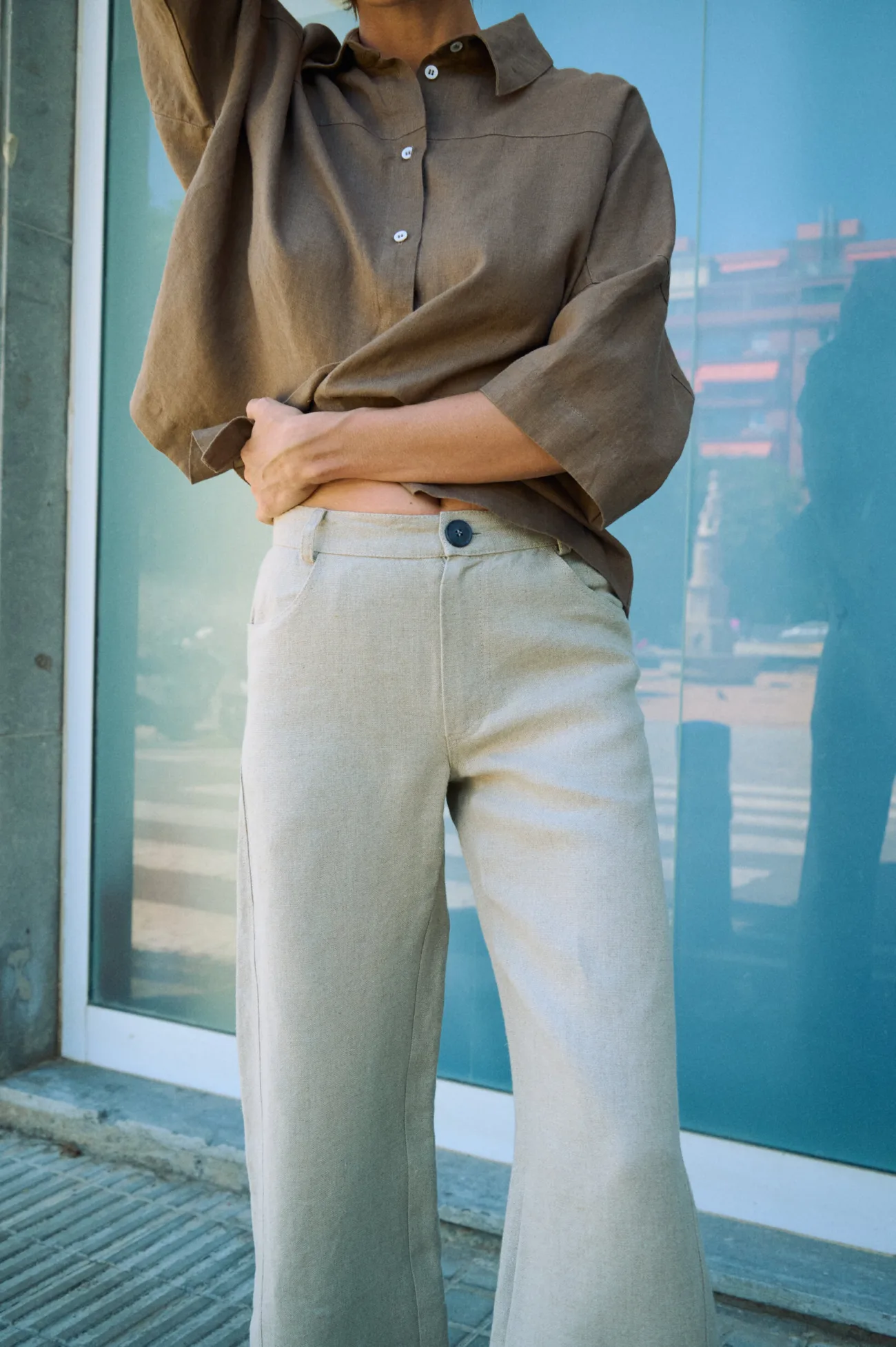 Natural grey heavy linen trousers with a tailored waistband, front button closure and wide-leg cut, paired with a brown linen shirt