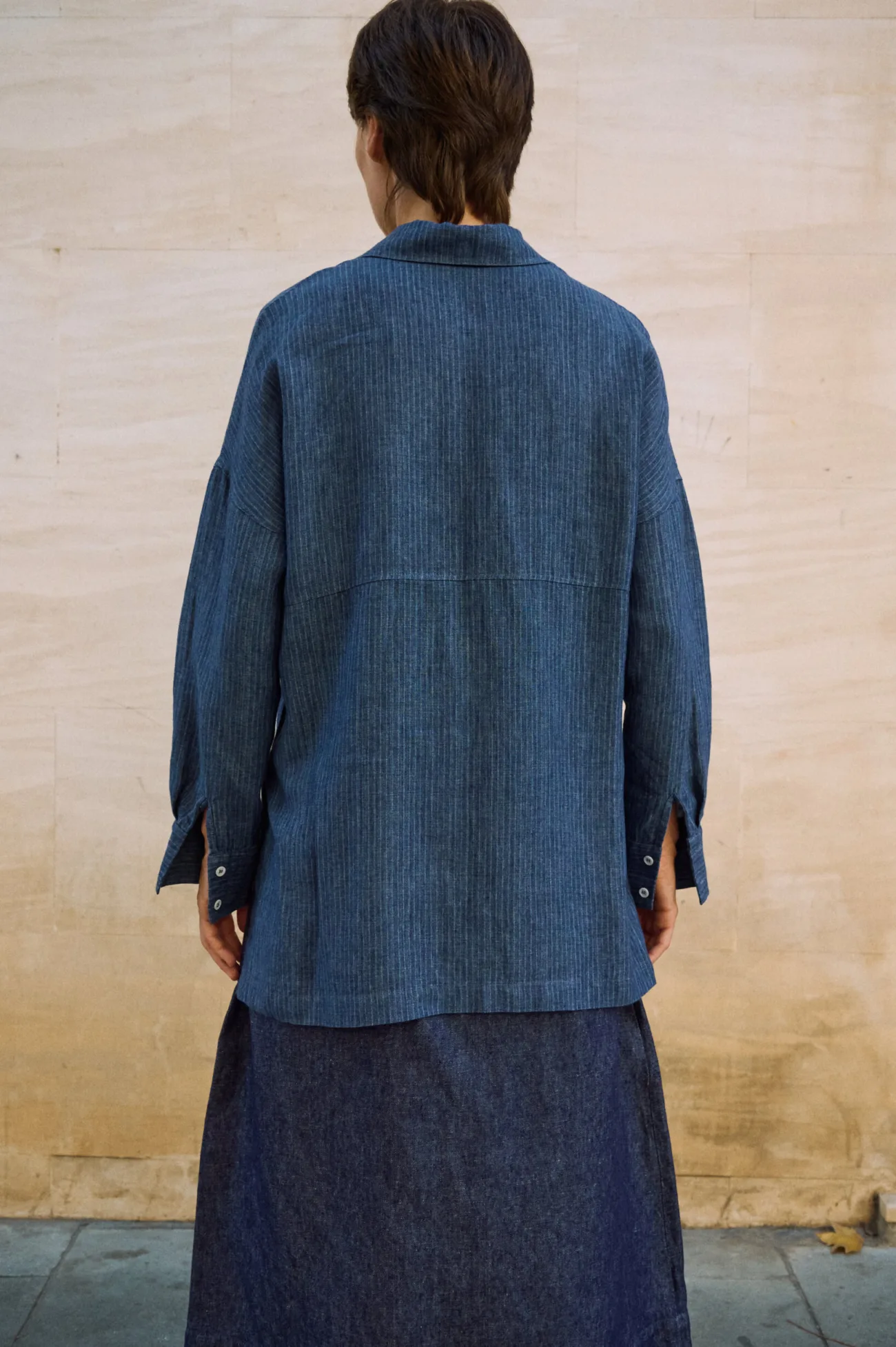 Back view of a blue striped linen button-up shirt with dropped shoulders, a horizontal back seam and long buttoned cuffs