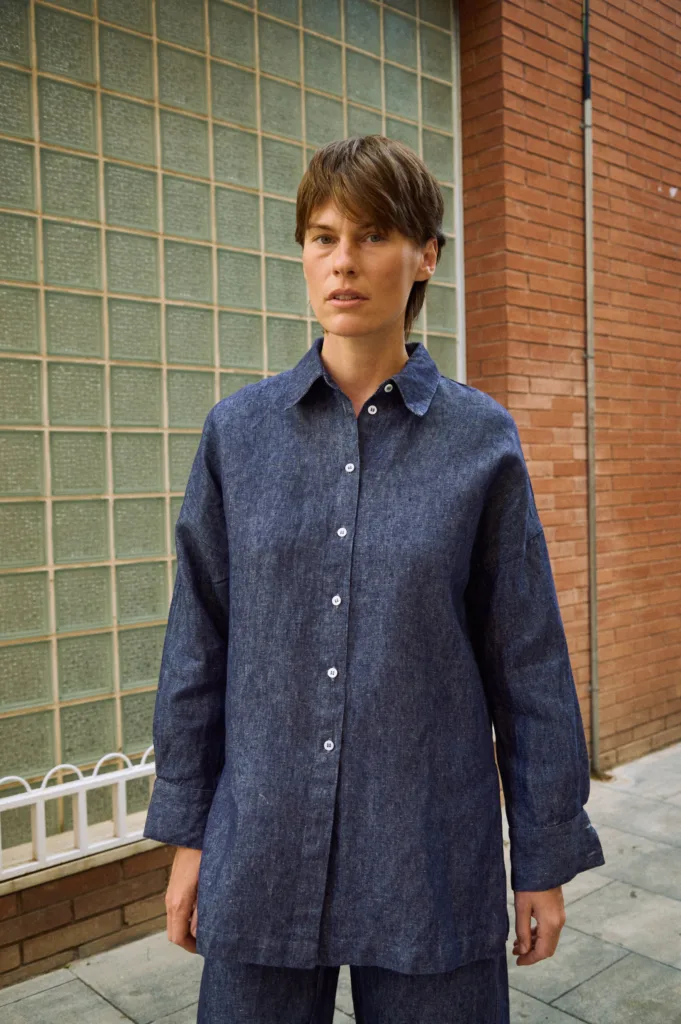 Front detail of blue denim linen shirt with button-down front, pointed collar, dropped shoulders, and wide cuffs