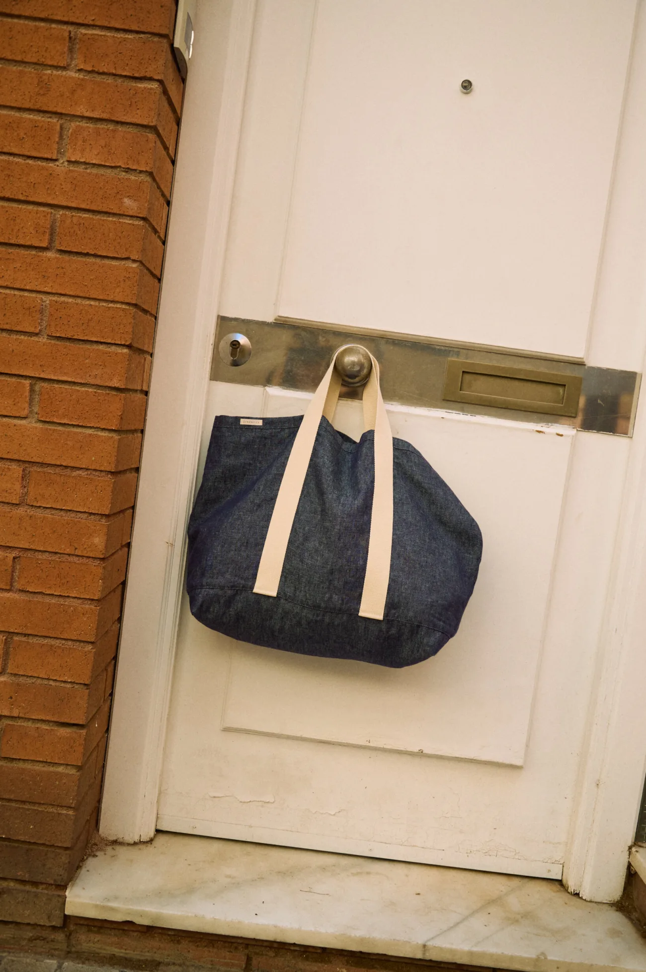 Large denim linen tote bag with wide cream cotton handles hanging against a white door
