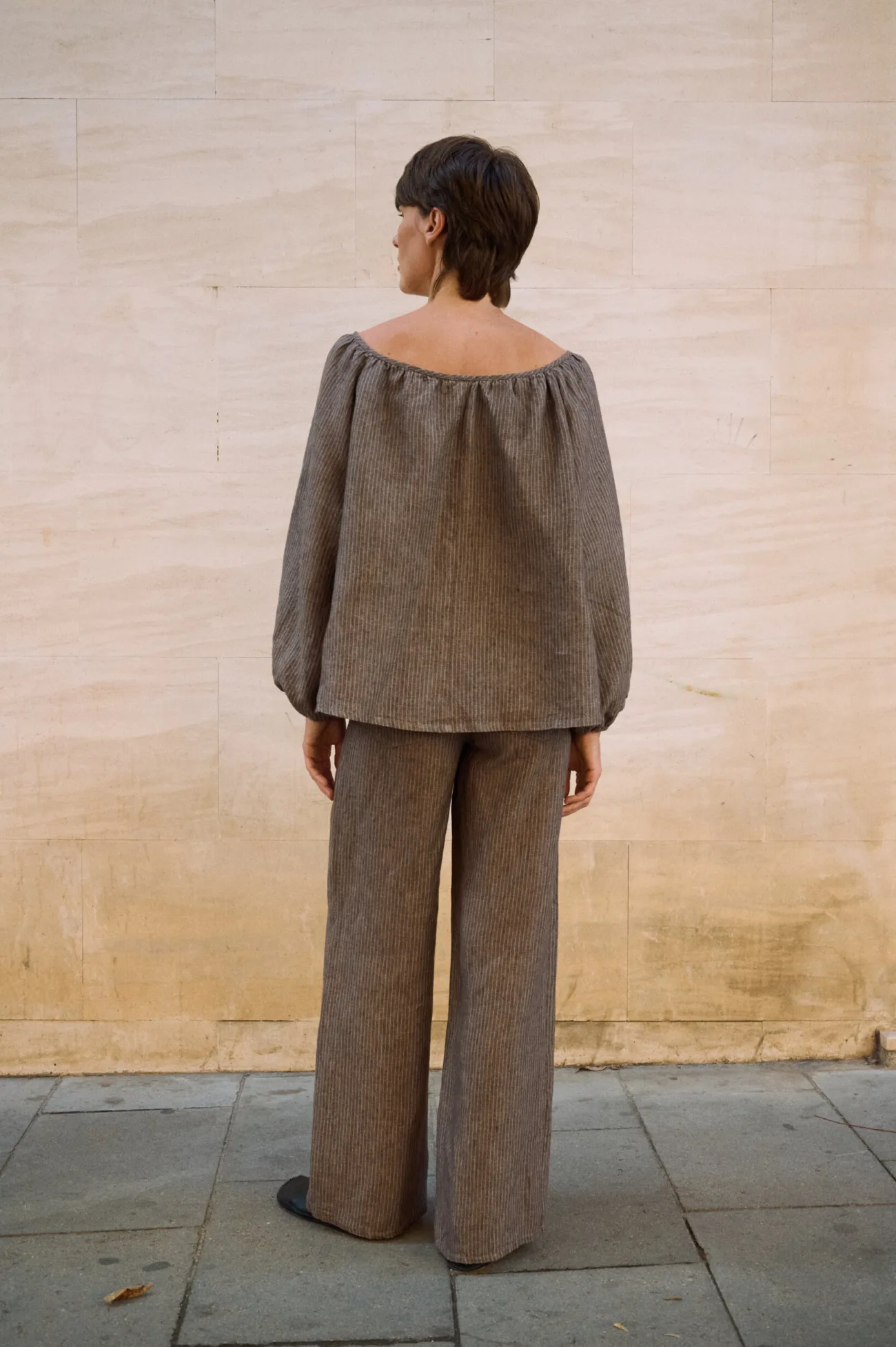 Back view of a brown striped linen blouse with a wide gathered neckline and voluminous sleeves, paired with matching wide-leg linen pants